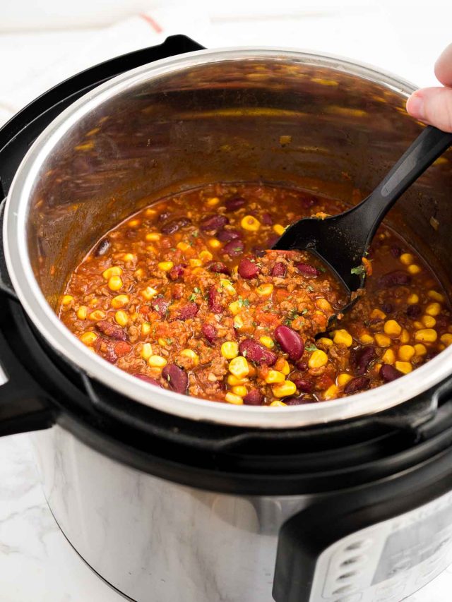 An instant pot containing chili con carne. A hand is holding a black cooking spoon in it.
