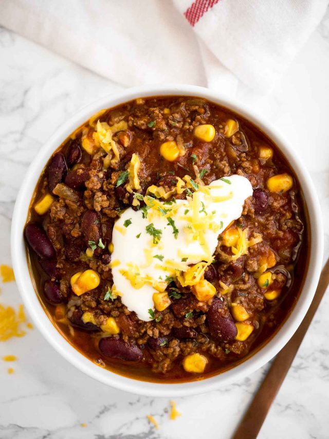 top-down shot of a white bowl of chili con carne, topped with sour cream, cilantro and shredded cheese on a marble surface with a white towel and a bronze spoon next to it.