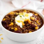 a white bowl of chili con carne, topped with sour cream, cilantro and shredded cheese on a marble surface with a white towel and a bronze spoon next to it.