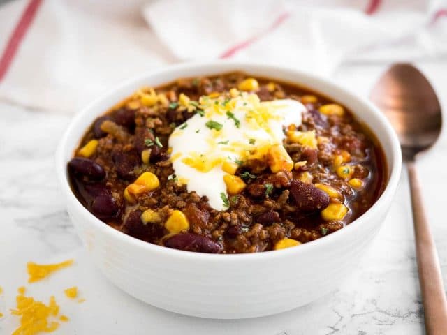 a white bowl of chili con carne, topped with sour cream, cilantro and shredded cheese on a marble surface with a white towel and a bronze spoon next to it.