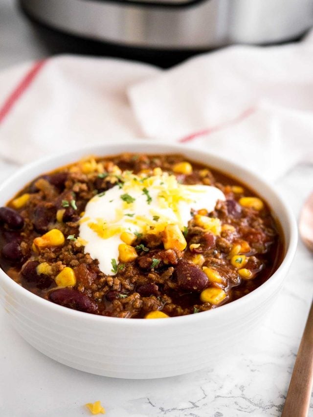 a white bowl of chili con carne, topped with sour cream, cilantro and shredded cheese on a marble surface with a white towel and a bronze spoon next to it.