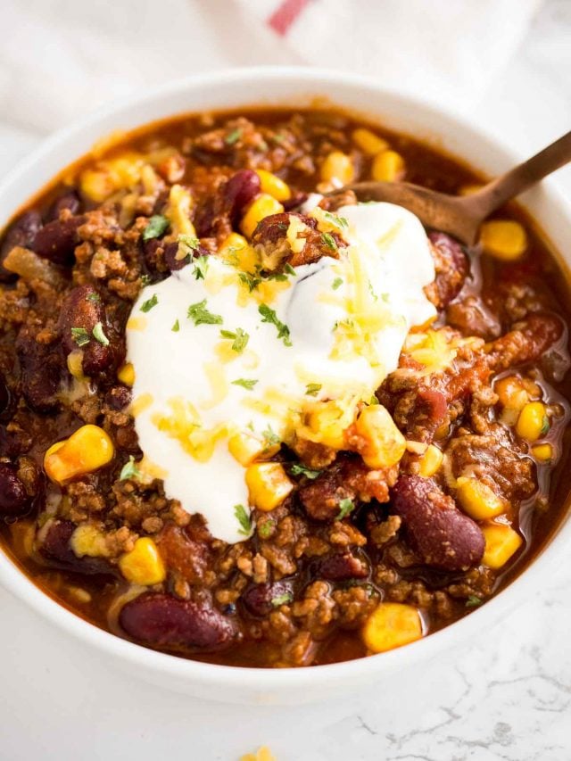 top-down shot of a white bowl of chili con carne, topped with sour cream, cilantro and shredded cheese with a bronze spoon in it.