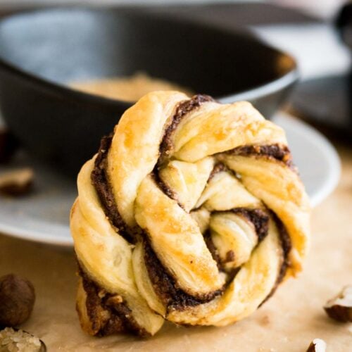 Close-up of a twisted nutella danish leaning against a black bowl with brown sugar garnished with hazelnuts on brown parchment paper.