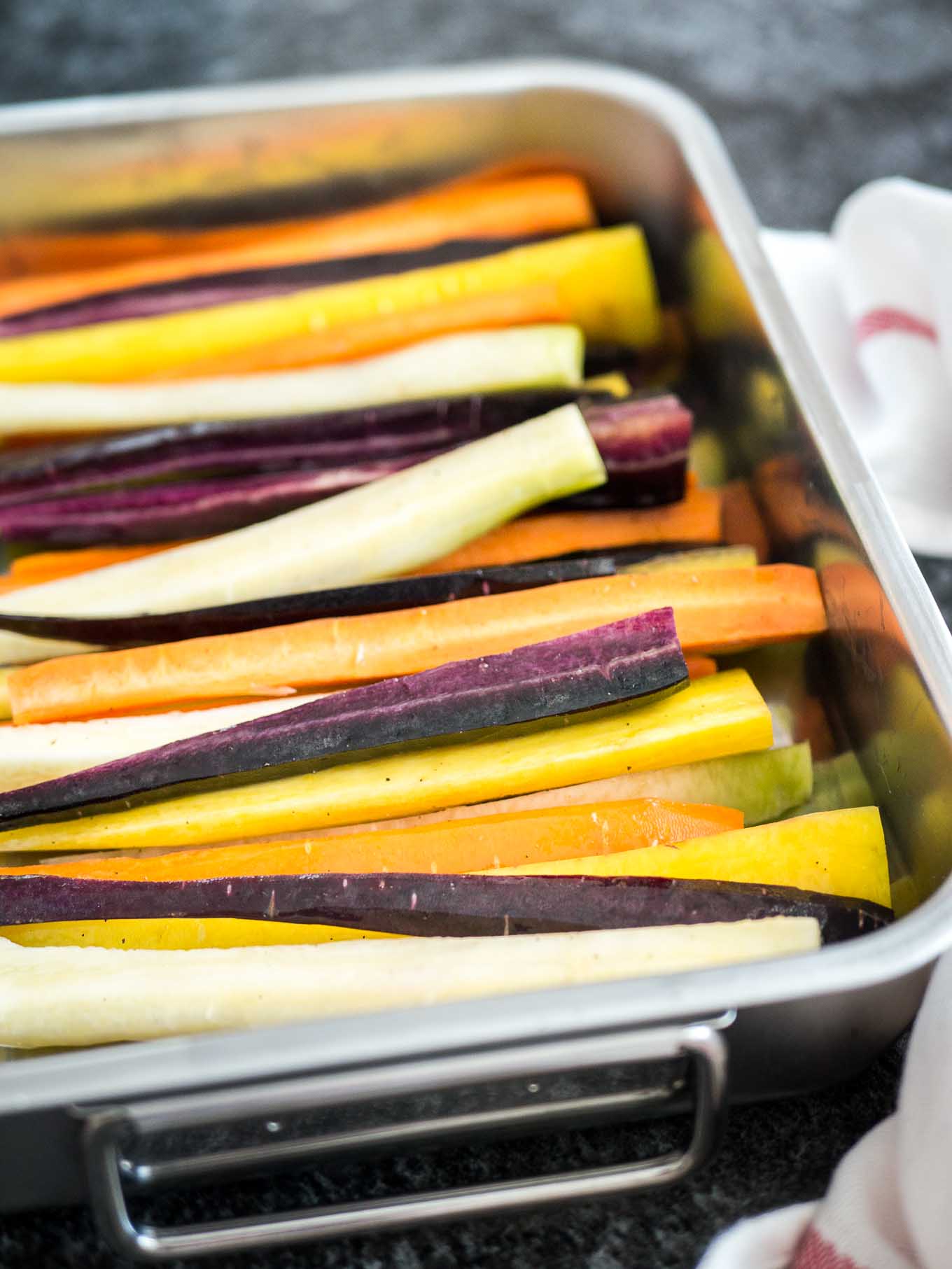 A roasting pan with rainbow carrots cut into wedges lengthwise before baking. There is a white dishtowel with a red stripe next to it.