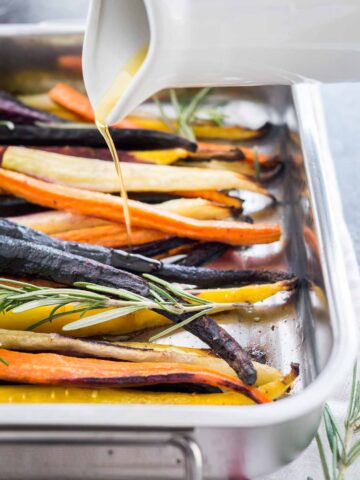 Close-up of a roasting pan with rainbow carrots garnished with sprigs of rosemary. A small white jug is pouring olive oil over it.