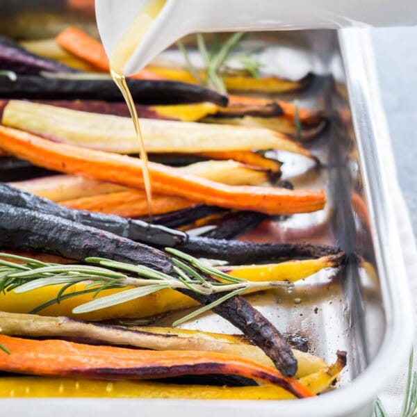 Close-up of a roasting pan with rainbow carrots garnished with sprigs of rosemary. A small white jug is pouring olive oil over it.