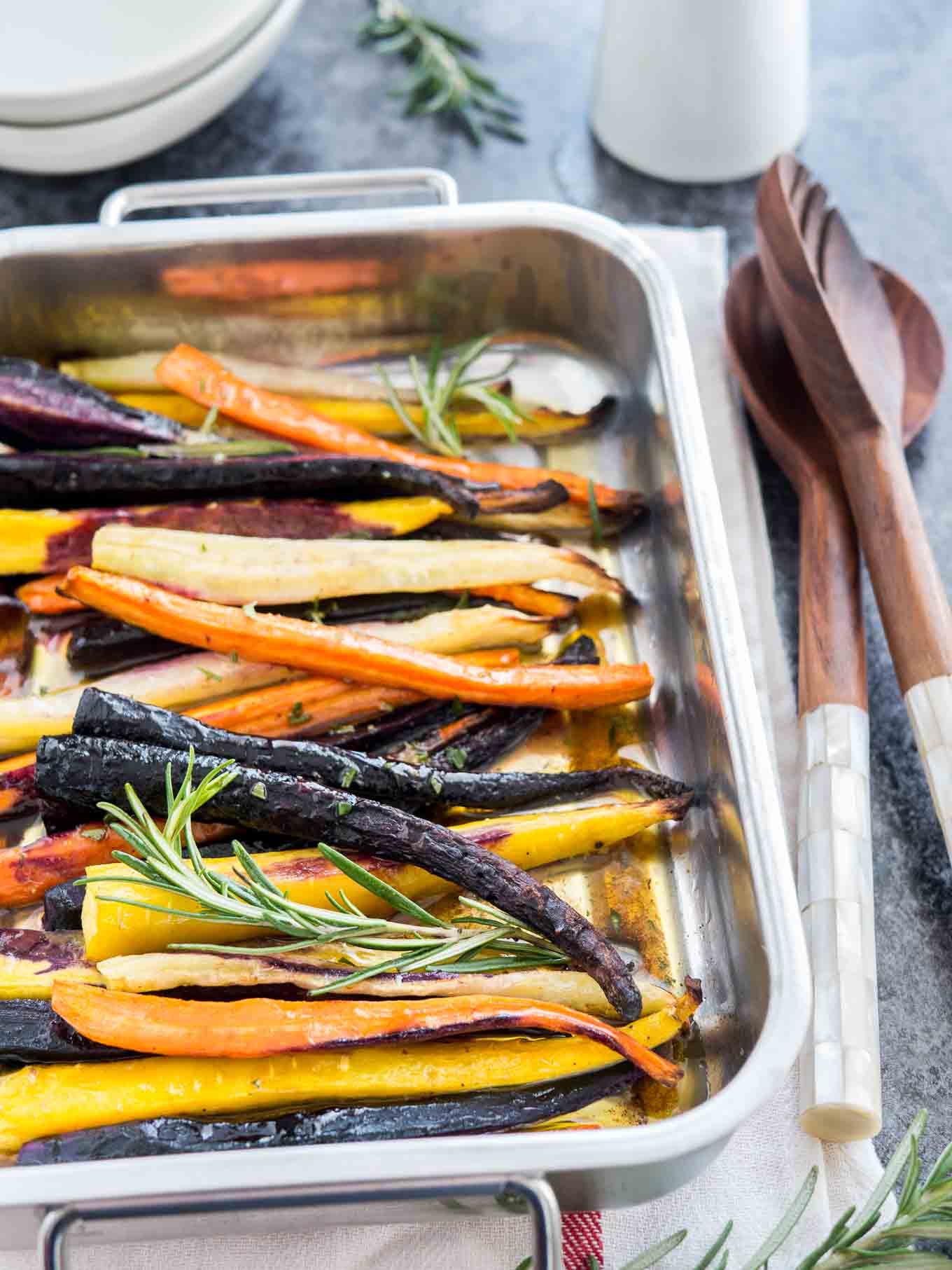 A roasting pan with roasted rainbow carrots garnished with sprigs of rosemary on a white dishtowel with a red stripe. There is a stack of plates in the background and salad tongs next to it.