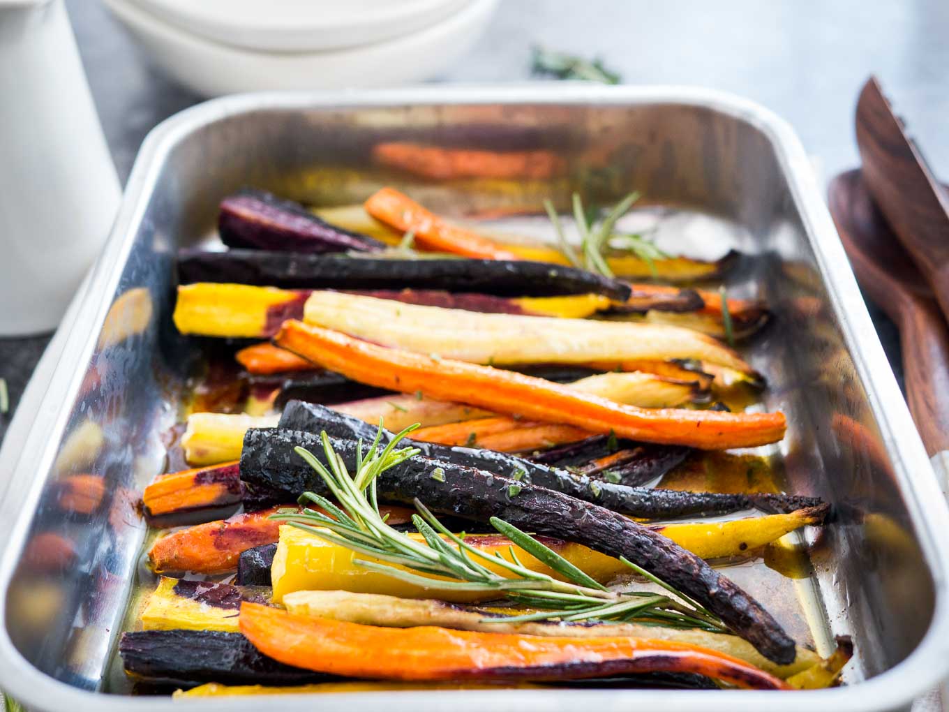 A roasting pan with roasted rainbow carrots garnished with sprigs of rosemary. There is a stack of plates in the background and salad tongs next to it.