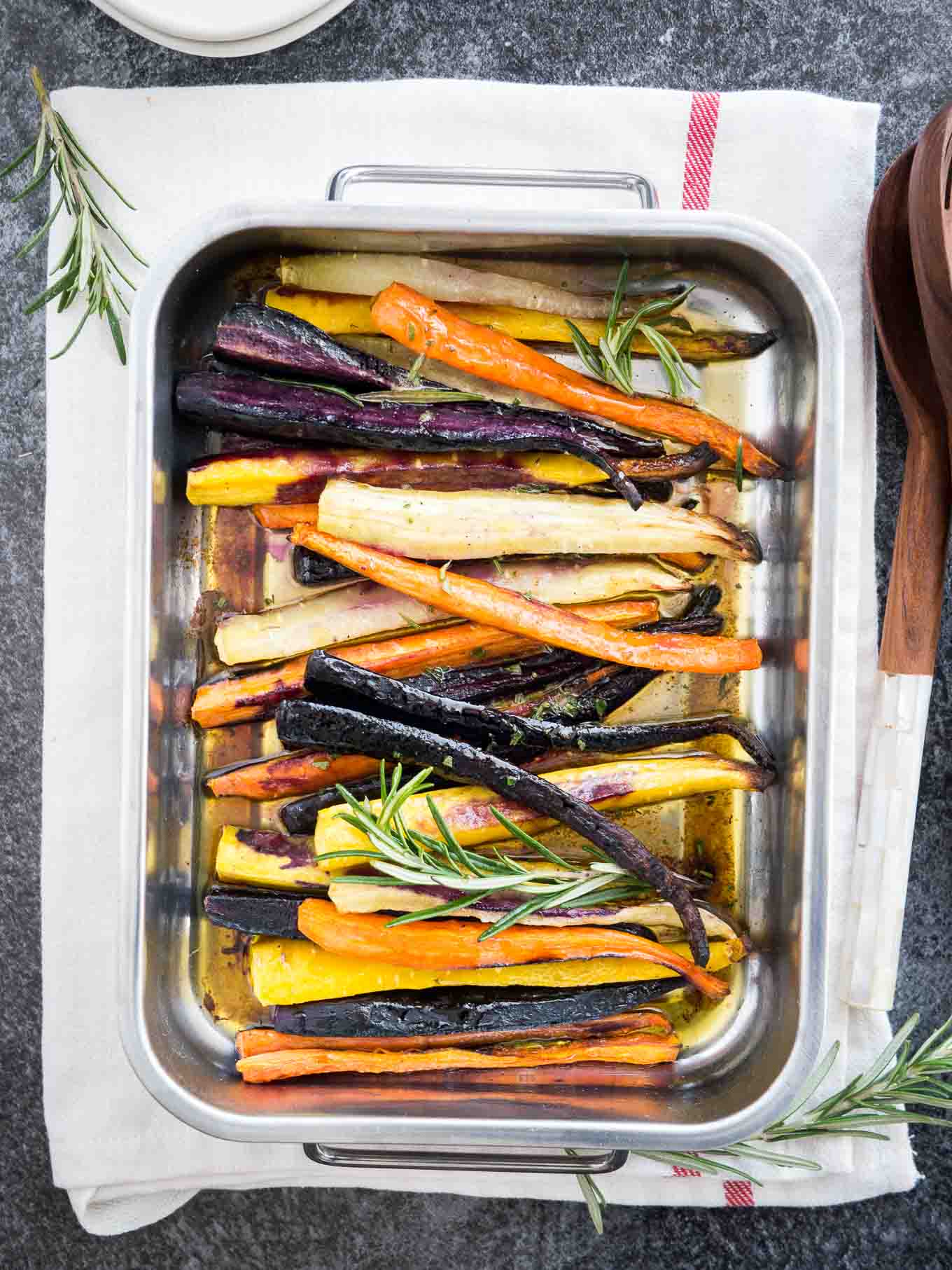 Top-down shot of a roasting pan with roasted rainbow carrots garnished with sprigs of rosemary on a white dishtowel with a red stripe. There are salad tongs next to it.
