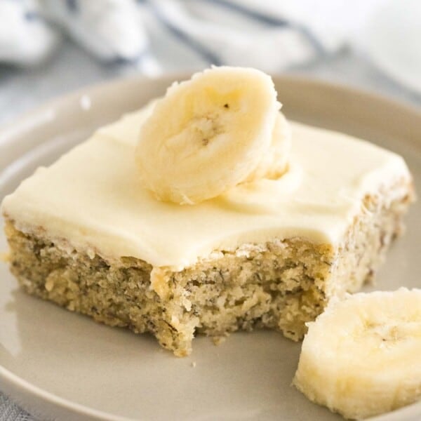 Close-up of a piece of banana cake with mascarpone frosting and topped with 2 banana slices on a grey plate on a grey tablecloth.  A bite has been taken out of the cake. There's a white and blue dishtowel in the background.