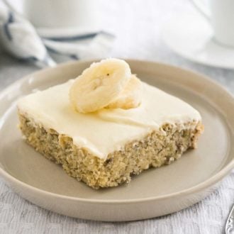 A piece of banana cake with mascarpone frosting and topped with 2 banana slices on a grey plate on a grey tablecloth. There's a fork next to it and a white cup in the background next to a white and blue dishtowel.