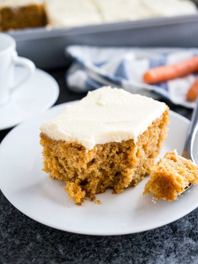 A piece of carrot cake with cream cheese frosting on a white plate. A piece has been taken out and is on a spoon next to it. There\'s a white and blue dishtowel in the background with 2 carrots on it.