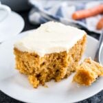 A piece of carrot cake with cream cheese frosting on a white plate. A piece has been taken out and is on a spoon next to it. There's a white and blue dishtowel in the background with 2 carrots on it.