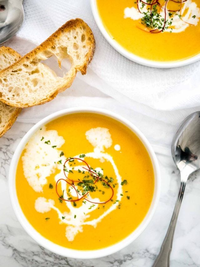 Top-down shot of two bowls of carrot soup with sour cream and chili threads with two toasted slices of bread and a spoon next to it on a white tablecloth.