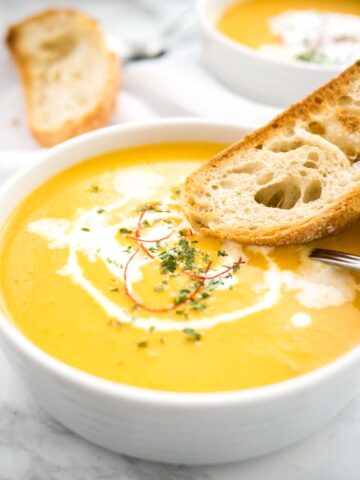 A bowl of carrot soup with sour cream and chili threads with a toasted slice of bread and a spoon in it. There's a toasted slice of bread and another bowl of soup in the background.