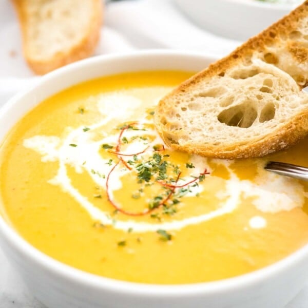 A bowl of carrot soup with sour cream and chili threads with a toasted slice of bread and a spoon in it. There's a toasted slice of bread and another bowl of soup in the background.