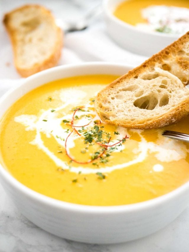 A bowl of carrot soup with sour cream and chili threads with a toasted slice of bread and a spoon in it. There\'s a toasted slice of bread and another bowl of soup in the background.