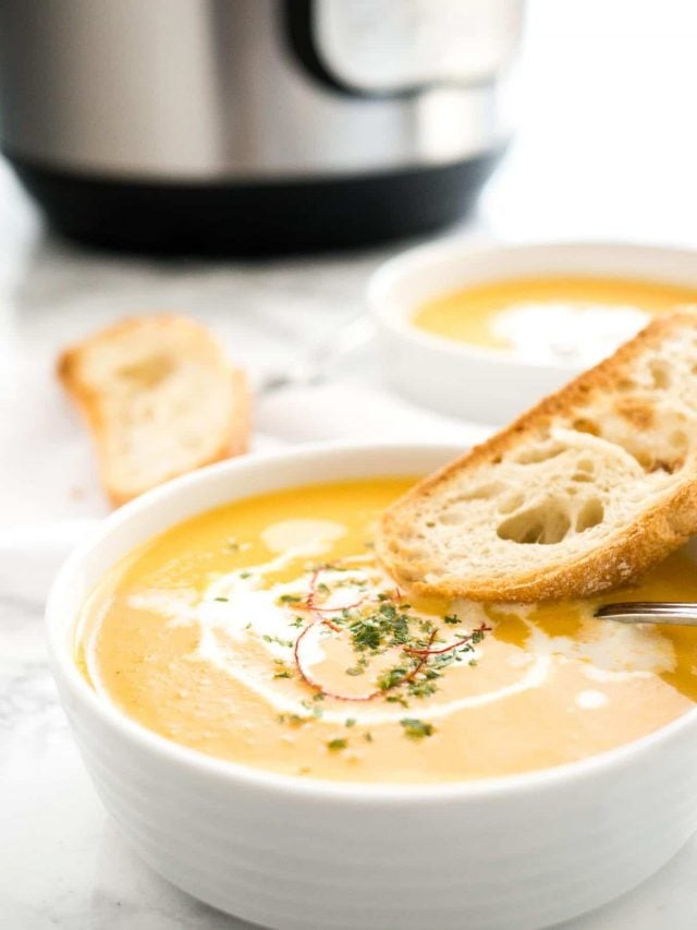 A bowl of carrot soup with sour cream and chili threads with a toasted slice of bread and a spoon in it. There\'s a toasted slice of bread, and instant pot and another bowl of soup in the background.