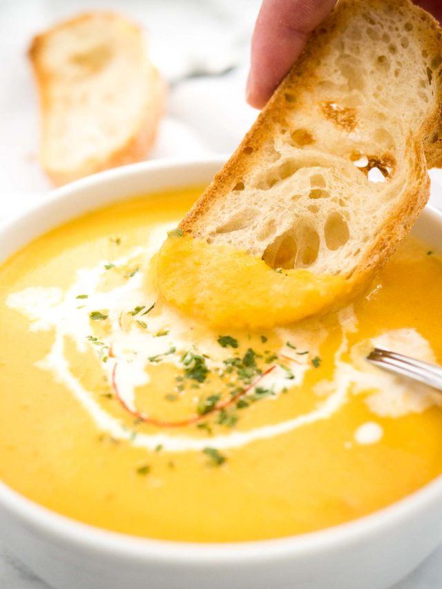 Close-up of a bowl of carrot soup with sour cream and chili threads with a spoon in it. A hand is dipping a toasted slice of bread into a soup. There\'s a toasted slice of bread in the background.