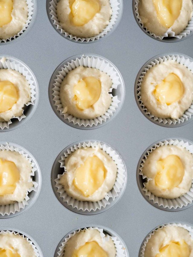 Top-down shot of a muffin pan, lined with muffin liners, filled with batter and dollops of lemon curd.