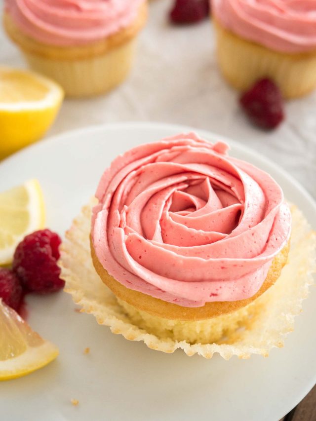 Close-up of a lemon raspberry cupcake with pink raspberry frosting, with the liner folded open on a white plate, garnished with lemon slices and raspberries. There are more of the cupcakes in the background.