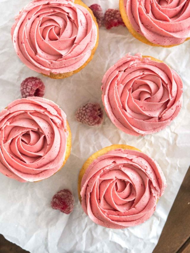 Top-down shot of raspberry lemon cupcakes with pink raspberry frosting next to some raspberries on parchment paper.