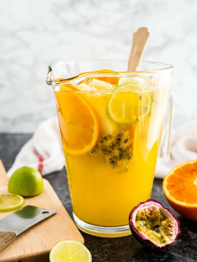 A glas pitcher or tropical white wine sangria with a wooden spoon, orange slices and passion fruit. There are limes, passion fruits and oranges next to it as well as a wooden cutting board and a white dishtowel.