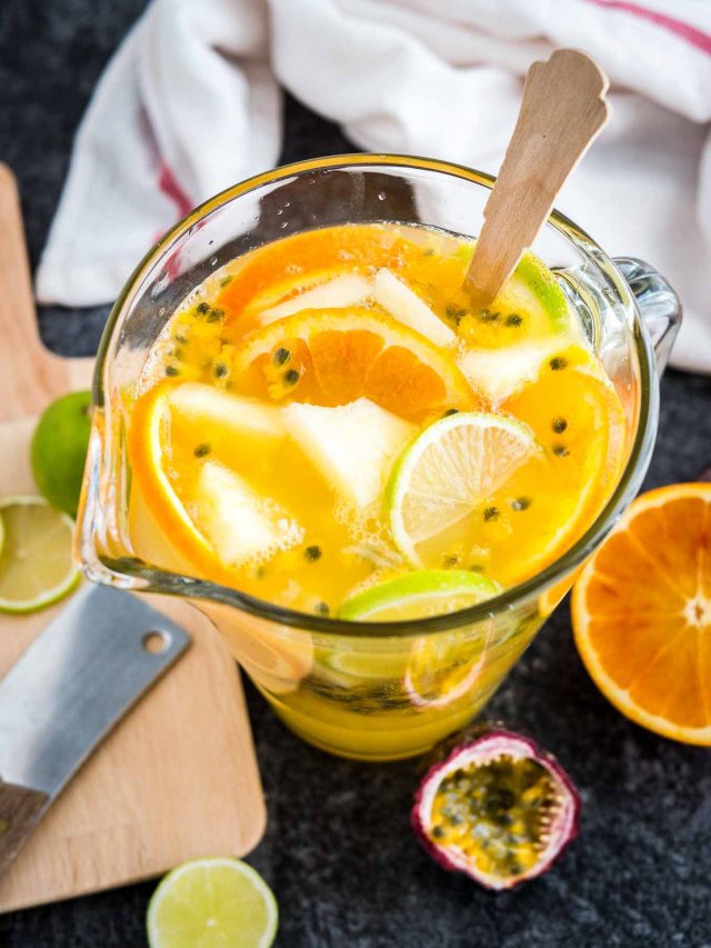 Top-down view of a glass pitcher of tropical white wine with orange slices and passion fruit next to a wooden cutting board and some fruit slices.