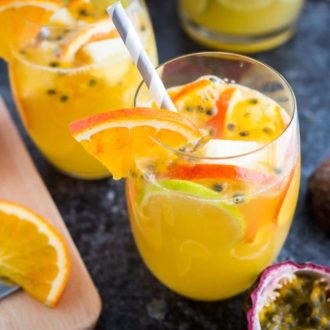 Two glasses of tropical white wine sangria with orange slices, passion fruit seeds and a straw on a dark surface. There are orange slices, another glass and passion fruits next to it.