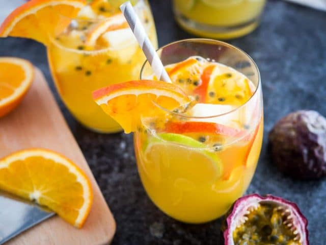 Two glasses of tropical white wine sangria with orange slices, passion fruit seeds and a straw on a dark surface. There are orange slices, another glass and passion fruits next to it.