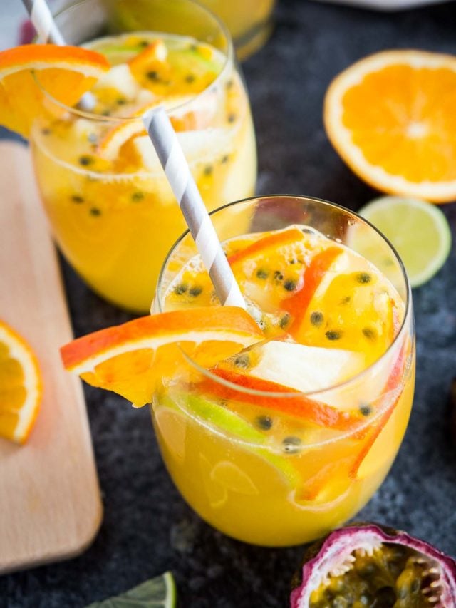 Top-down shot of a glass of tropical white wine sangria with orange slices, passion fruit seeds and a straw on a dark surface. There are orange slices, another glass and passion fruits next to it.