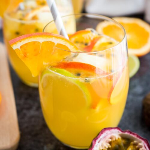 Close-up of a glass of tropical white wine sangria with orange slices, passion fruit seeds and a straw on a dark surface. There are orange slices, another glass and passion fruits next to it.