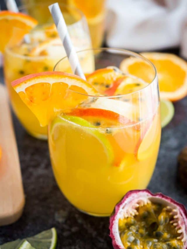 Close-up of a glass of tropical white wine sangria with orange slices, passion fruit seeds and a straw on a dark surface. There are orange slices, another glass and passion fruits next to it.