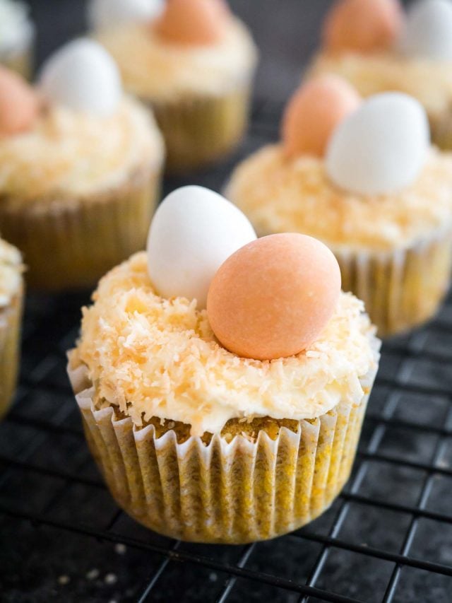 Carrot cake cupcakes, topped with cream cheese frosting, coconut and Cadbury sugar eggs, sitting on a black cooling rack