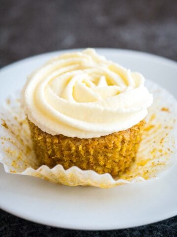 A carrot cake cupcake with cream cheese frosting on top. The paper is folded away from the cupcake and it's sitting on a white plate