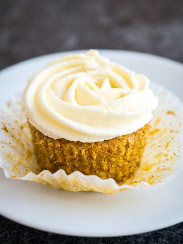  Un cupcake de pastel de zanahoria con glaseado de queso crema en la parte superior. El papel se pliega lejos de la magdalena y se sienta en un plato blanco