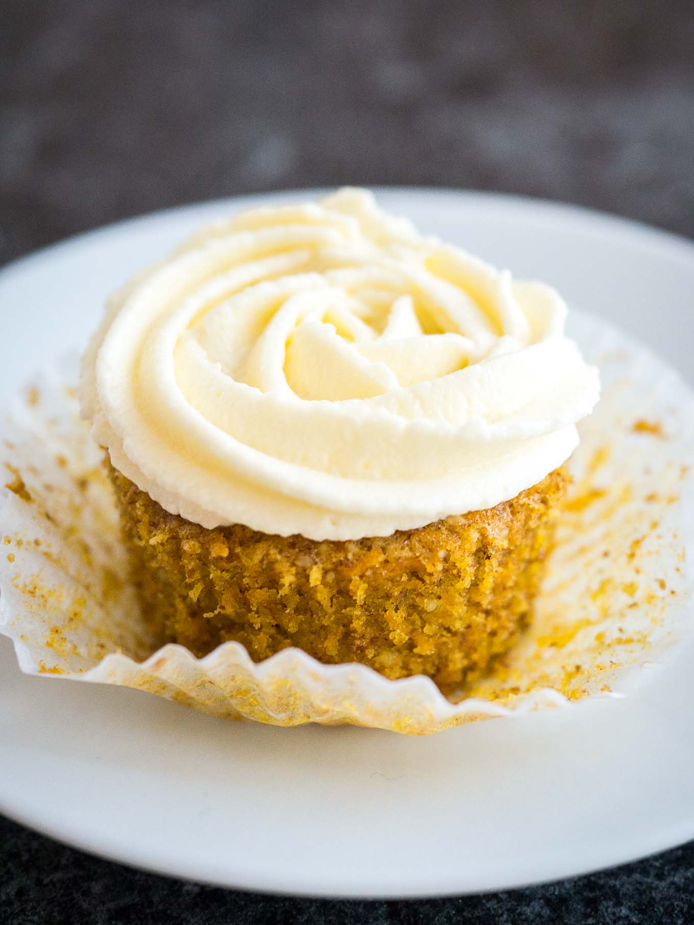 A carrot cake cupcake with cream cheese frosting on top. The paper is folded away from the cupcake and it's sitting on a white plate.