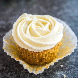 A carrot cake cupcake with cream cheese frosting on top. The paper is folded away from the cupcake and it's sitting on a dark grey surface.