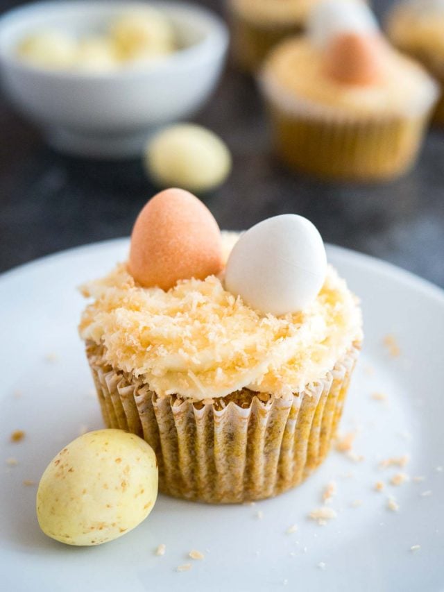 Carrot cake cupcake, topped with cream cheese frosting, coconut and Cadbury sugar eggs, sitting on a white plate next to a sugar egg.
