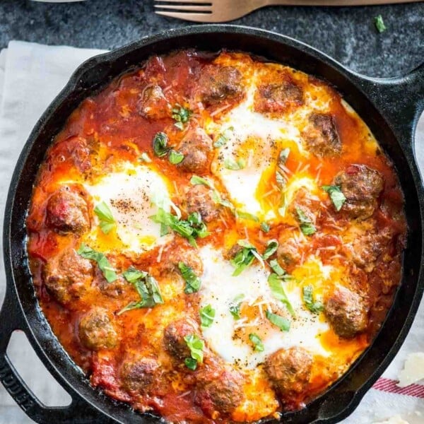 Top-down shot of a cast iron pan of Italian baked eggs and meatballs garnished with parsley on a white and red dishtowel. There's a stack of white plates, a bronze fork and some pieces of Parmiggiano next to it.