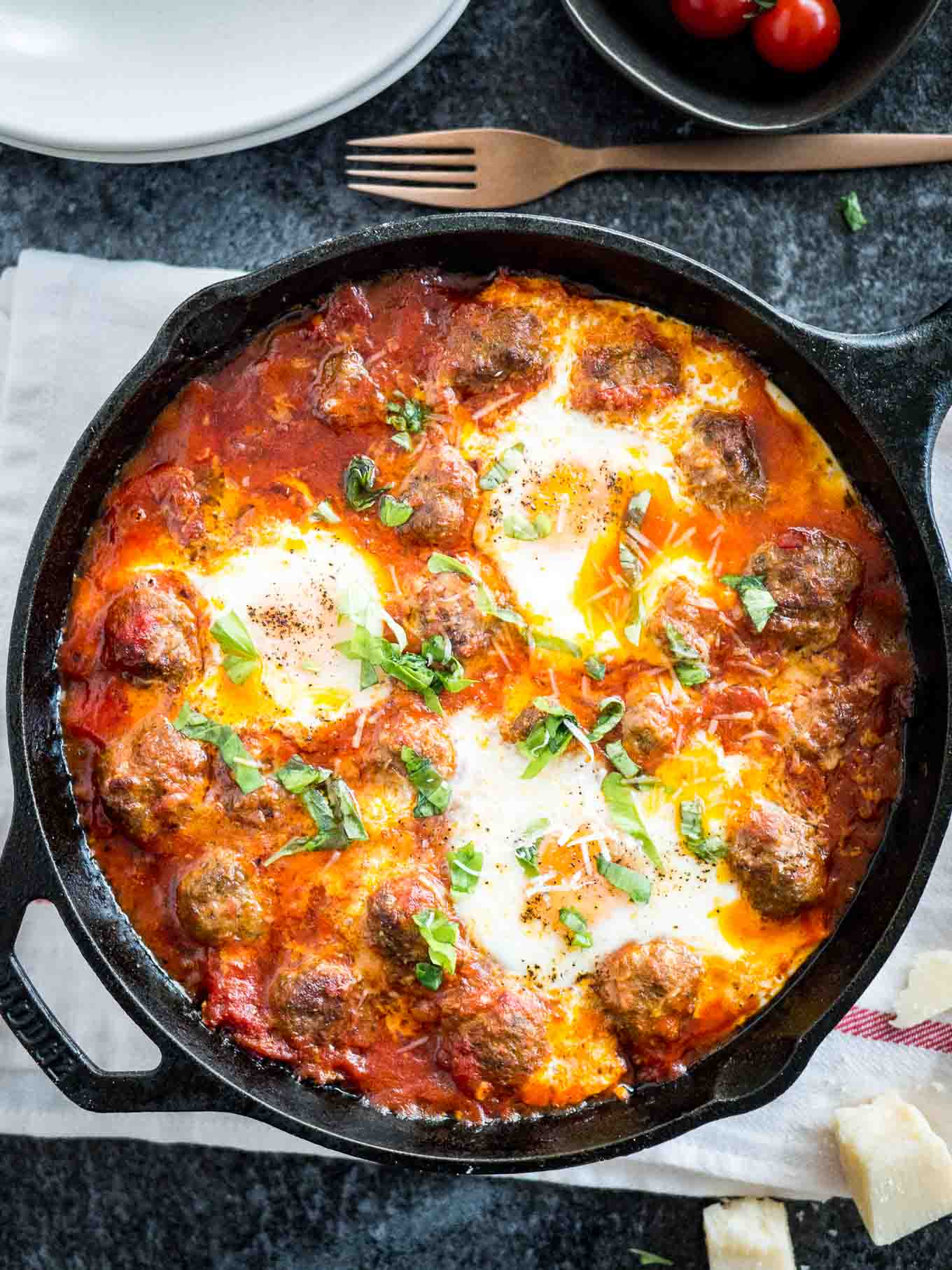 Top-down shot of a cast iron pan of Italian baked eggs and meatballs garnished with parsley on a white and red dishtowel. There\'s a stack of white plates, a bronze fork and some pieces of Parmiggiano next to it.