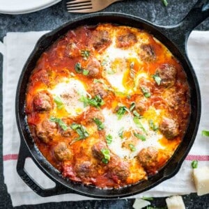 Top-down shot of a cast iron pan of Italian baked eggs and meatballs garnished with parsley on a white and red dishtowel. There's a stack of white plates, a bronze fork and some pieces of Parmiggiano next to it.