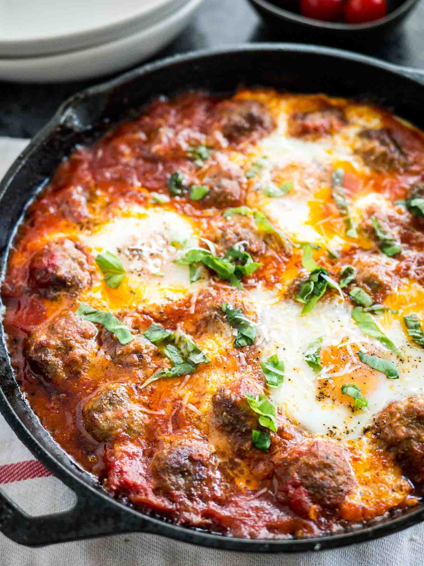 Close-up shot of Italian baked eggs and meatballs garnished with parsley in a cast iron pan on a white and red dishtowel.