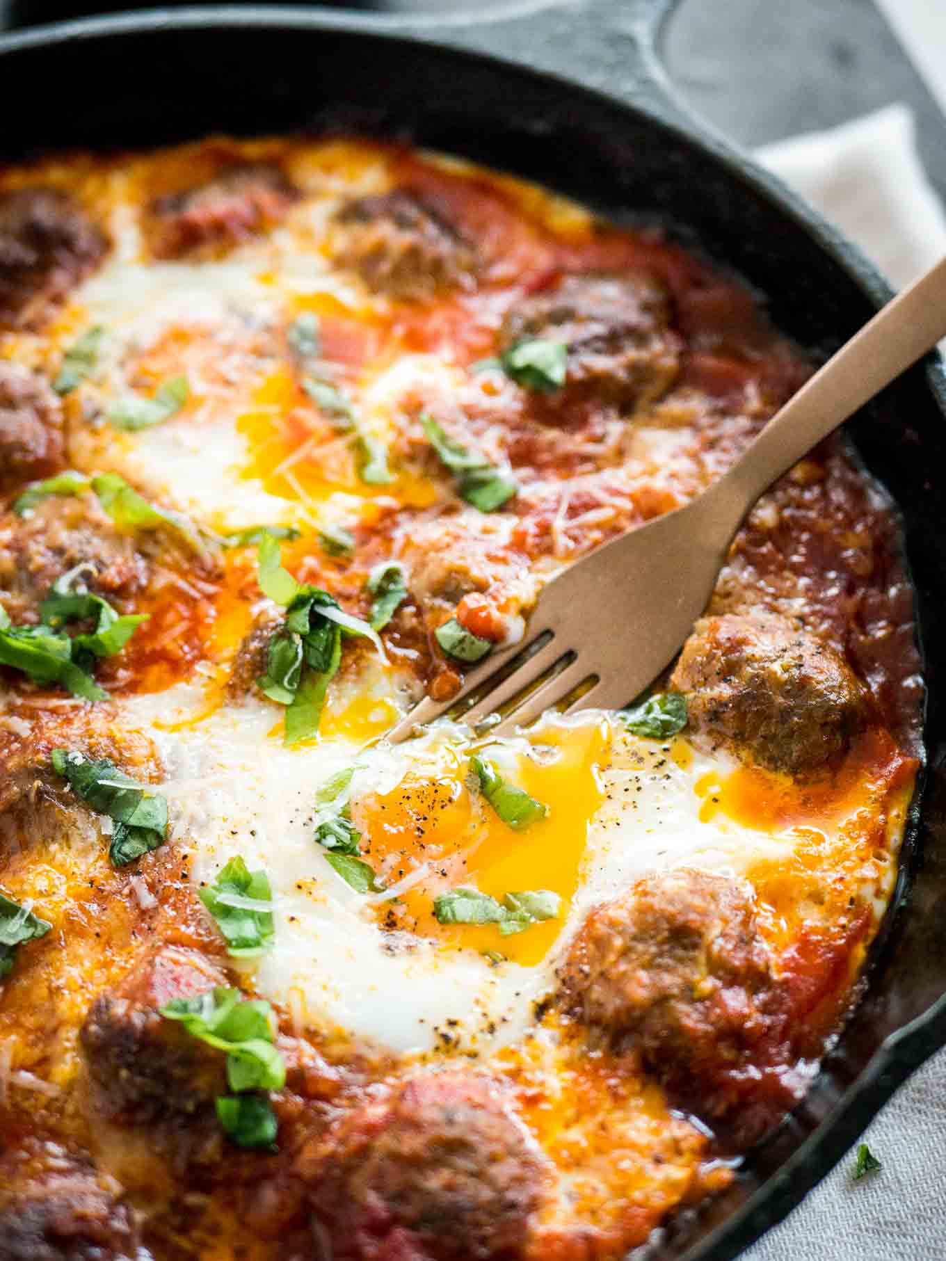 Close-up shot of Italian baked eggs and meatballs garnished with parsley in a cast iron pan with a bronze fork.