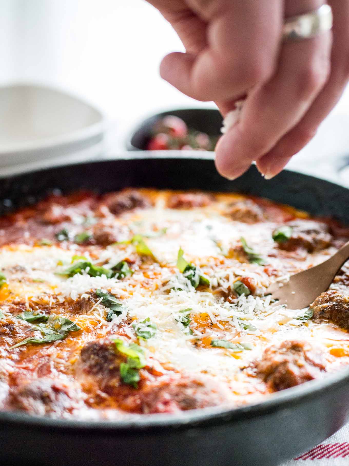A hand crumbling Parmiggiano onto a cast iron pan of Italian baked eggs and meatballs with a bronze fork in it.