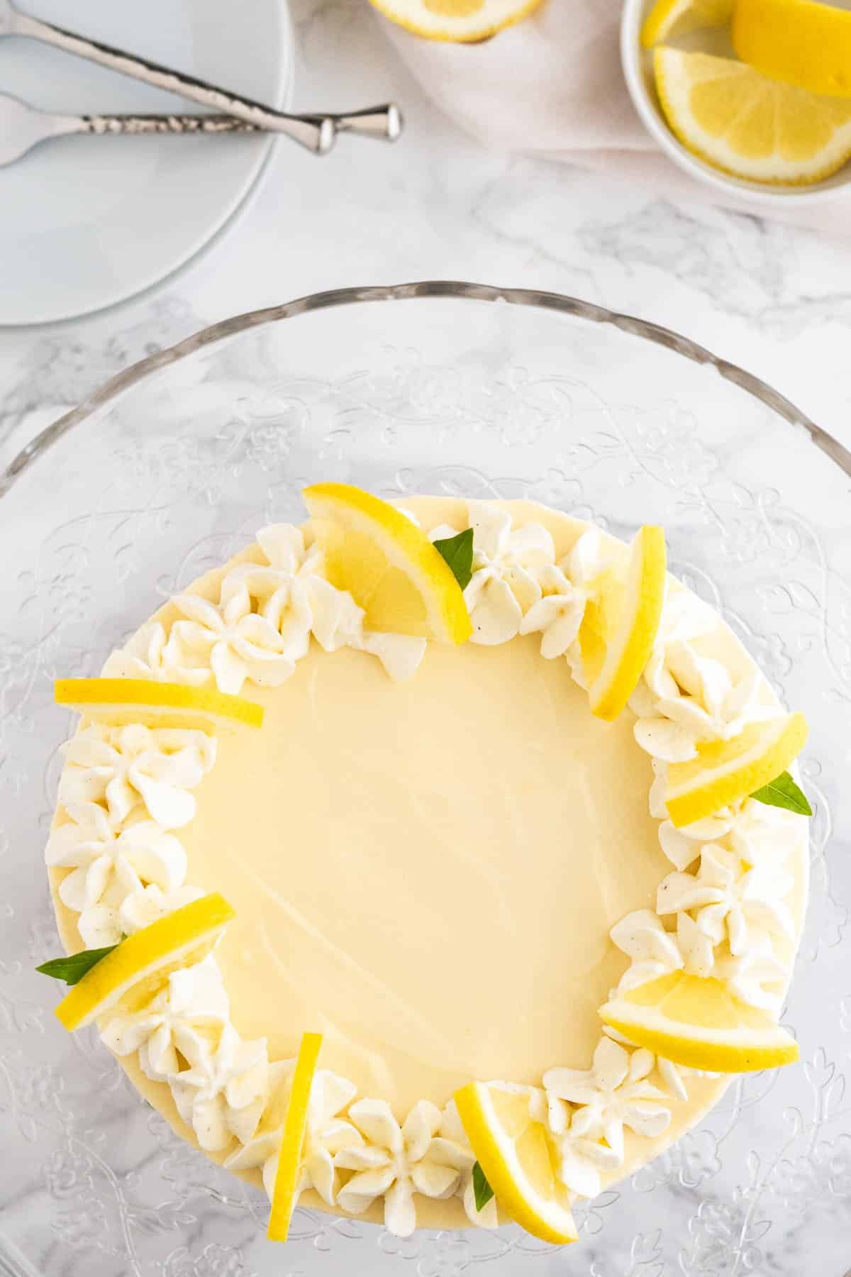 Top-down shot of a lemon cream pie topped with whipped cream and lemon slices on a glass serving platter on a marble surface. 