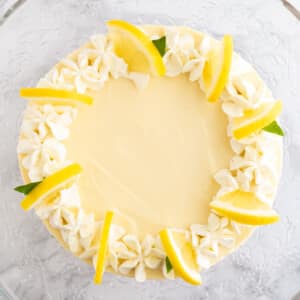 Top-down shot of a lemon cream pie topped with whipped cream and lemon slices on a glass serving platter on a marble surface.