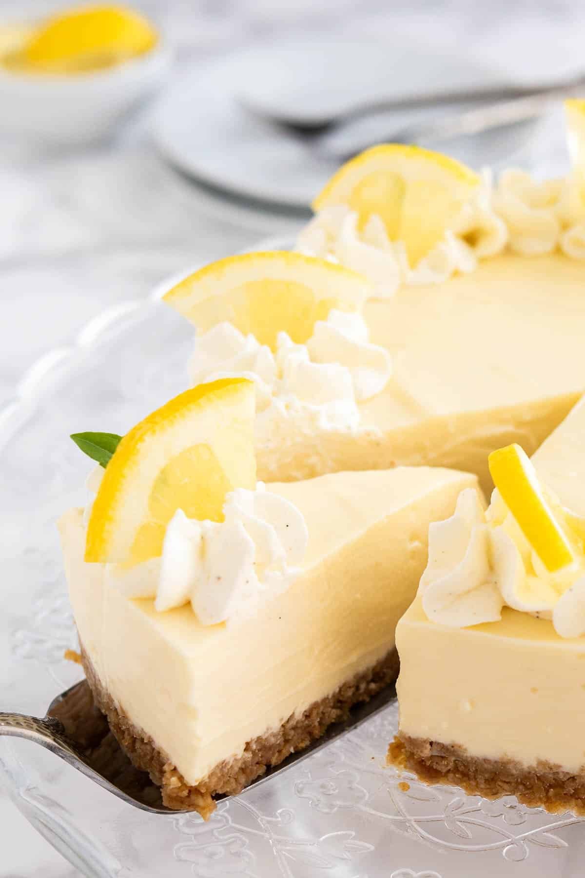 A lemon cream pie on a glass serving platter topped with whipped cream and lemon wedges. A cake server is lifting out a slice.