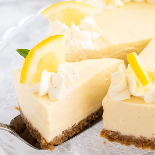 A lemon cream pie on a glass serving platter topped with whipped cream and lemon wedges. A cake server is lifting out a slice.