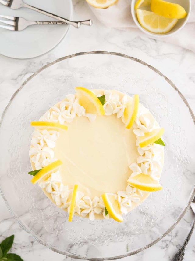 Top-down shot of a lemon cream pie topped with whipped cream and lemon slices on a glass serving platter on a marble surface. There\'s a stack of white plates with forks and a small white bowl of lemon slices next to it.